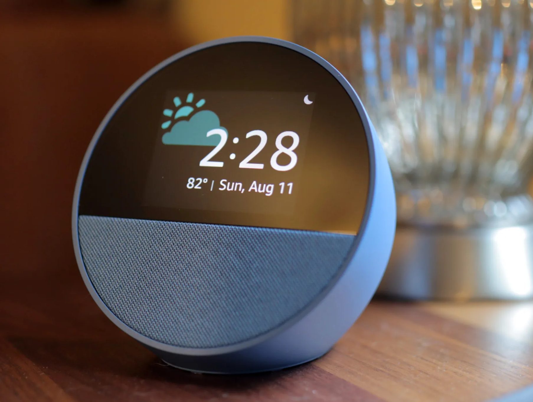 Blue Amazon Echo Spot on a wooden surface with a silver lamp in the background.